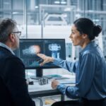 Zwei Geschäftsleute in einem modernen Büro diskutieren vor einer Glaswand mit Haftnotizen, ein Laptop zeigt eine KI-Datenanalyse. Die Szene symbolisiert die Verbindung von menschlicher Kommunikation und Technologie im Consulting